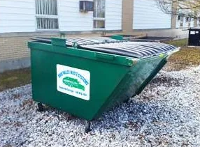 A green dumpster sitting in the gravel outside of a house.